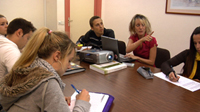 Imagen de una Imagen de una reunión de empresa en la que aparecen varias personas sentadas alrededor de una mesa. Todos están tomando apuntes a excepción de una mujer que, junto a un proyector, explica algo a sus compañeros.