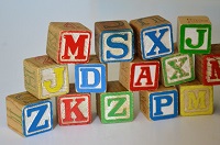 EIlustración en la que se ve un juego de bloques de madera con cubos que forman parte de un puzzle en tres dimensiones. Los cubos contienen el abecedario en letras mayúsculas y minúsculas así como algunos dibujos con letras.