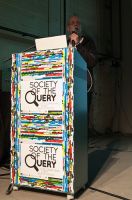 Fotografía de Escenario que tiene un atril forrado de carteles en los que puede leerse “Society of Query”, en castellano significa “La sociedad de la consulta”.