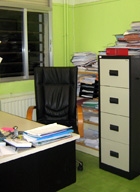 Puesto de trabajo en el que se aprecia una mesa con algunos papeles, una silla ergonómica y detrás un gran archivador y una estantería llena de carpetas.