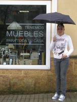 Leticia, la joven  propietaria de una tienda de muebles, posa delante del escaparate de su tienda. Sujeta un paraguas abierto porque el día está lluvioso.