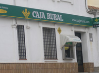 Fotografía de la fachada de una entidad financiera, Caja Rural. Se observa el letrero verde y el logotipo característico de la entidad; y un cajero automático, cubierto con un toldo blanco junto a la puerta.