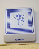 Cartel colgado en la pared en cuya base se
indica la leyenda “Silencio” en el que sobre fondo blanco hay unas líneas azules que enmarcan los labios,
aparentemente de una mujer, que se lleva el dedo índice hacia ellos para indicar silencio.