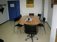 Día a día. Imagen de una mesa de reuniones dentro de un despacho. Se aprecia una puerta al fondo de color azul, una mesa ovalada de madera y seis sillas negras rodeándola.