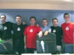 En la imagen se muestran a seis hombres, compañeros de trabajo de pie frente a la cámara, dos de ellos visten una camiseta roja y el resto camiseta negra. Todos están alegres y sonrientes.