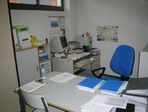 Oficina 2. En la foto se ve un despacho vacío, con la silla de color azul y el mobiliario en blanco. Hay distinta documentación encima de la mesa.