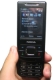 En la imagen se ve en primer plano una mano que sujeta un teléfono móvil de color negro. En la pantalla del mismo se pueden ver distintos caracteres de las utilidades.