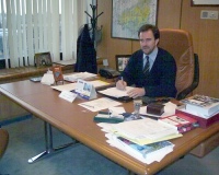 Se observa a un directivo con traje de color oscuro en su mesa de despacho con gran cantidad de documentación sobre ella.