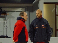 Conversación. En la imagen se muestra a dos hombres de mediana edad, uno vestido con cazadora roja y negra y otro con cazadora negra que están hablando en un local comercial.