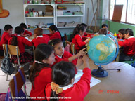Grupo. En la imagen se observa varios escolares con uniforme rojo, que trabajan en pequeños grupos en el aula. Un grupo está manejando una bola del mundo.