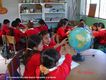En la imagen se observa varios escolares con uniforme rojo, que trabajan en pequeños grupos en el aula. Un grupo está manejando una bola del mundo.