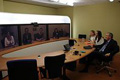 Tres pantallas en la pared, donde se reflejan personas dialogando con las que estan sentadas en la mesa de la la sala.