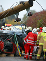 Se ve un poste de un tendido eléctrico caído en primer plano, y detrás, dos coches accidentados, uno encima de otro, y varias personas con vestimentas de equipos de emergencia  alrededor.