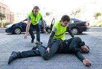 Una víctima está tendida en el suelo. Un miembro del equipo de emergencias acaba de ponerle en posición lateral de seguridad. Otra mujer del equipo de emergencias se acerca apresurada hasta la víctima para ayudar en su atención. Ambos miembros del equipo de emergencias visten chalecos reflectantes de seguridad.