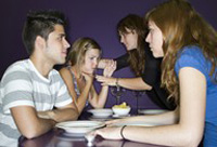 Cuatro jóvenes sentados en una mesa de restaurante. Chico joven sentado en un restaurante frente a una muchacha. A su lado otra chica se lleva las manos al cuello. Frente a ella, una chica de pelo oscuro se inclina a ayudarla desde el otro lado de la mesa.