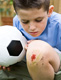 Fotografía de un niño de unos siete años con una pelota de fútbol. Está agachado mirándose una pequeña herida en la rodilla. Hay restos de hierba alrededor de la herida por lo que parece producida por una caída mientras jugaba..