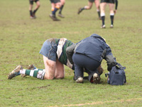 Fotografía de un campo de fútbol en el que un jugador está arrodillado en el suelo aparentemente lesionado. Un técnico sanitario con un botiquín portátil está agachado a su lado intentando ayudarle.