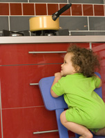 Fotografía de un niño muy pequeño que está subido a una silla intentando alcanzar un cazo situado en el fuego de la hornilla. El riesgo de volcarse el cazo caliente encima es muy evidente en la imagen.