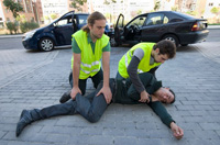 Imagen de dos personas socorriendo a un hombre tumbado en el suelo. El herido está colocado lateralmente y los socorristas están arrodillados a su lado uno sujetándole las piernas y el otro mirando atentamente el rostro del herido.