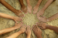 Fotografía en la que aparece un grupo de manos con las palmas hacia abajo, de personas de distintas razas, colocadas formando un círculo sobre la arena de una playa.