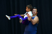 Fotografía de 2 bailarines del psico ballet de Maite León que recoge un momento de la coreografía en el que un hombre de mediana edad, con Síndrome de Down, eleva en brazos a un niño que realiza una figura artística con las piernas abiertas flexionadas en ángulo recto respecto a su cuerpo.