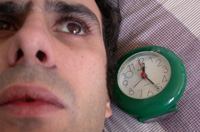 Fotografía de un hombre en cama junto a un reloj despertador con gesto de tener dificultades para dormir.