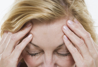 Fotografía del rostro de una mujer con los ojos cerrados y las manos en la cabeza con gesto de sentir molestia física o dolor.