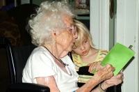 Foto apaisada en la que se ve una abuela sobre su silla de ruedas y al lado de pie junto a ella, una niña pequeña rubia, de unos tres años. Ambas miran un libro de portada verde.