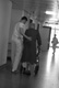  Foto en blanco y negro, en la que se aprecia en el pasillo de un hospital, a un joven con su uniforme sanitario y a un paciente anciano al que le va ayudando en su deambulación. 