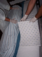 En la imagen podemos ver como un técnico sujeta al paciente que está en decúbito lateral dentro de la cama, bajo la encimera. Otro técnico, estira la bajera limpia en la orilla de la cama contraria.