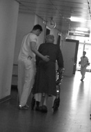 Un técnico, vestido con el pijama sanitario, acompaña por el pasillo a un anciano ayundándolo en su deambulación.