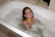 Niña pequeña sonriente aparece en una bañera llena con espuma y utilizando una esponja amarilla.