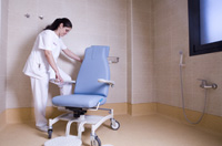 Mujer morena, con pelo oscuro recogido y uniforme blanco en cuarto de baño preparando silla adaptada para el aseo.