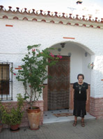 María de 79 años posa de cuerpo entero delante de su casa, tiene el pelo corto, teñido de negro. Viste falda y camiseta negras con guirnaldas de flores pintadas por ella misma. Calza sandalias, al cuello tiene el colgante blanco con el botón rojo de teleasistencia. Detrás la fachada de su casa, una casa mata con un tejado de tejas rojas, la pared es blanca y la puerta principal está tapada con una cortina exterior de colgantes de madera. A la derecha de María se ven varias macetas y parte de una ventana con reja.