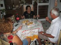 En la sala de María. Sentados en torno a una mesa redonda en la que se ven pasteles, copas y refrescos se ve a tres personas, de frente María de 79 años tiene el pelo corto, teñido de negro. Viste camiseta negra y al cuello tiene el colgante blanco con el botón rojo de teleasistencia. Frente a ella y de perfil, casi de espaldas a la Cámara están Chelo con el pelo castaño corto y rizado y camiseta estampada y Tomás con Camisa blanca y el pelo muy blanco, son matrimonio y conviven juntos.