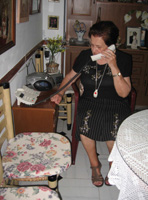 En la sala de María. Ella está sentada en una silla hablando por el teléfono que se encuentra en una mesilla a su derecha. Tiene 79 años, el pelo corto, teñido de negro. Viste falda y camiseta negras con guirnaldas de flores pintadas por ella misma. Calza sandalias, al cuello tiene el colgante blanco con el botón rojo de teleasistencia.