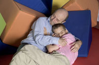 En un fondo de colchonetas de colores (parece una sala de rehabilitación psicomotriz). Recostado en una de ellas, un padre calvo, con barba de varios días y camisa a rayas, abraza a su hijo paralítico cerebral sentado sobre el. Es un joven adolescente moreno con camiseta rosa y un pantalón claro.