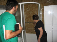 En el baño de María. Dos personajes de perfil. María ha abierto la mampara de la ducha y la muestra a Leonardo.  Leonardo observa con atención lo que María le señala en sus manos sostiene un bloc de notas. María de 79 años tiene el pelo corto, teñido de negro. Viste camiseta y falda negras.  Leonardo: Hombre de 38 años, moreno con barba y bigote, viste camiseta verde.