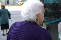 Primer plano del perfil de una anciana que está en la calle, apenas se le ve la cara, tiene el pelo muy blanco y un pendiente pequeño en la oreja que se le ve, viste una rebeca morada. Al fondo otra anciana con una muleta y vestida con chaqueta verde y falda negra, cruza la calle.