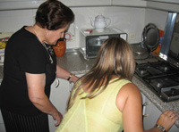 En la cocina de María. Dos personas se inclinan a mirar el interior de un mueble bajo. María, de perfil, mira al mueble que se encuentra debajo de la hornilla, está un poco inclinada hacia  adelante y señalando algo. Esther, a su lado, de espaldas a la cámara, se agacha y observa con atención lo que María le señala. María de 79 años tiene el pelo corto, teñido de negro. Viste camiseta y falda negras y al cuello tiene el colgante blanco con el botón rojo de teleasistencia. Esther de unos 24 años, rubia con el pelo muy largo lleva un vestido verde claro.