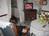 Fotografía de la salita de María. De frente puede verse un mueble con la televisión encendida, en primer plano y de espaldas al espectador, María está sentada en su butaca viendo la televisión, a penas se le ve el pelo corto y moreno y un brazo con una manga negra, a su derecha hay una mesa camilla.