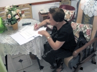 Plano de cuerpo entero de María. Una mujer de 79 años. Está sentada en la butaca de su salita y se inclina porque está escribiendo sobre una mesa en una hoja de papel, sostiene entre las manos un bolígrafo, está muy atenta a su labor. Le gusta mucho arreglarse y ponerse guapa, tiene el cabello corto, teñido de negro  Viste camiseta y falda negras.