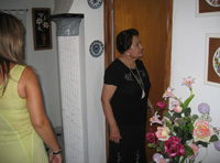 María de perfil abriendo una puerta para entrar en una habitación, mientras Esther está dirigiéndose hacia ella de espaldas a la cámara. María mujer de 72 años vestida de negro y Esther de 22 con vestido verde.