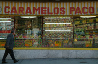 Escaparate de una tienda de caramelos, donde se ve una gran variedad y en la calle un hombre pasa caminando y mirando atentamente los caramelos. Encima hay un tolodo a rayas amarillas y blancas con la inscripción Caramelos Paco.