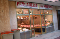 Fachada de una pescadería. Puede verse el escaparate y al otro lado los expositores con el pescado y un pescadero. Encima del escaparate hay un panel con la leyenda Pescadería La morena, fuera en la calle hay dos contenedores de basura pequeños.