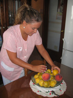 Esther 22 años, rubia con el pelo recogido en una coleta rizada y uniforme de TAPD a rayas rosas y blancas. De pie en la cocina está inclinada sobre un frutero colocando melocotones y ciruelas.