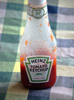 Bote de ketchup marca Heinz, transparente de plástico al que le queda poco tomate y está colocado sobre un mantel de cuadros azules y blancos.