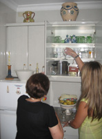 En la cocina de María todo el mobiliario es blanco. María (72 años, pelo pintado de castaño, vestida de negro) y Esther (22 años, rubia, pelo largo, vestida de verde) están de espaldas a la cámara, la chica colocando unas copas de cristal en un estante de un mueble que permanece abierto.