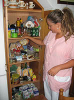 Junto a una despensa (armario de madera de gran tamaño) con la puerta abierta, llena de productos alimenticios no perecederos de todo tipo, dispuestos en baldas. Esther (22 años, rubia, pelo recogido en una coleta. Con uniforme de TAPD de rayas rosas y blancas) se dispone a ordenarla.