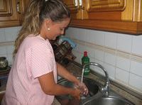 Esther (22 años rubia de pelo largo y recogido en una coleta, con uniforme de rayas rosas) está en la cocina enjuagando un vaso bajo un chorro de agua en el fregadero.